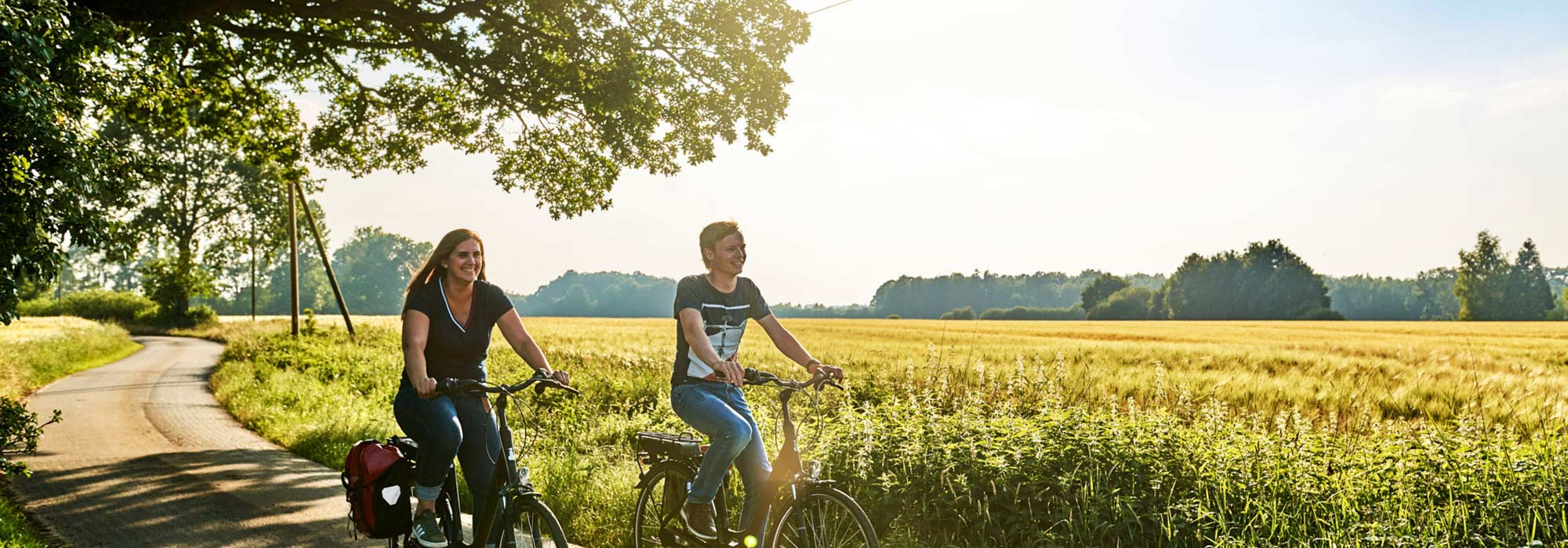 Ein Mann und eine Frau fahren mit dem Rad bei sommerlichem Wetter einen Weg zwischen Wiesen und Feldern