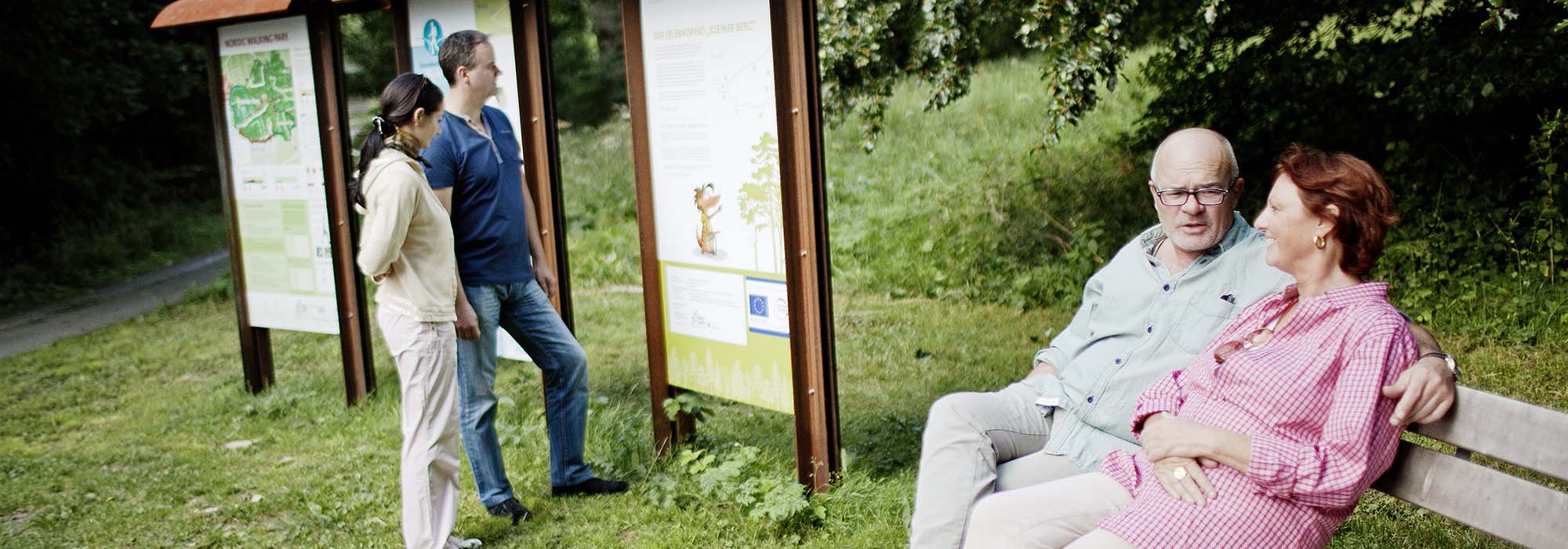 Ein Mann und eine Frau sitzen auf einer Bank im Grünen, während zwei andere Menschen sich eine Informationstafel ansehen