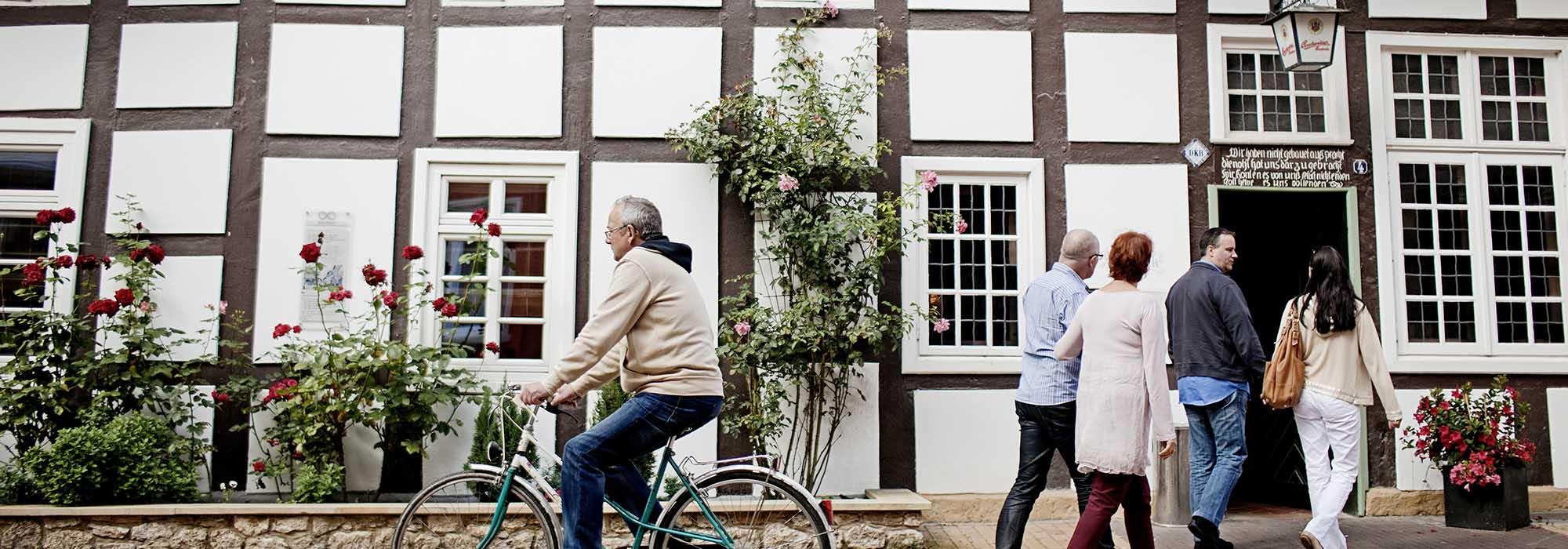 Ein Mann fährt mit einem Fahrrad vor einem Fachwerkhaus her, eine kleine Menschengruppe geht in die entgegengesetzte Richtung