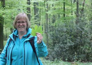 Eine Frau streckt lächelnd ein Ahornblatt im Wald in die Kamera.