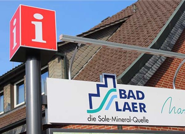 Informationszeichen und ein Schild mit dem Bad Laer Logo bei blauem Himmel und Haus im Hintergrund