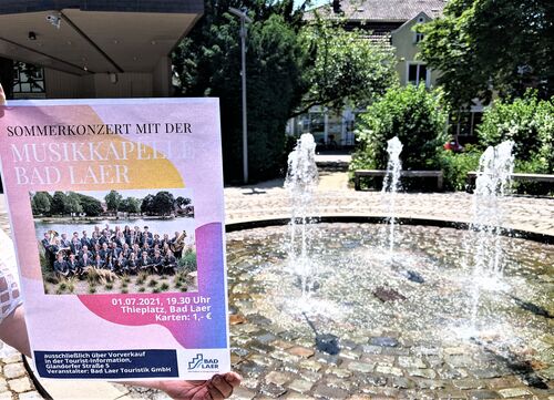 Plakat zum Sommerkonzert der Musikkapelle Bad Laer wird vor Konzertmuschel und Brunnen am Thieplatz gehalten.