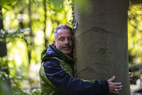 Ein Mann mittleren Alters umarmt lächelnd mit geschlossenen Augen einen Baum.