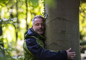 Ein Mann mittleren Alters umarmt lächelnd mit geschlossenen Augen einen Baum.