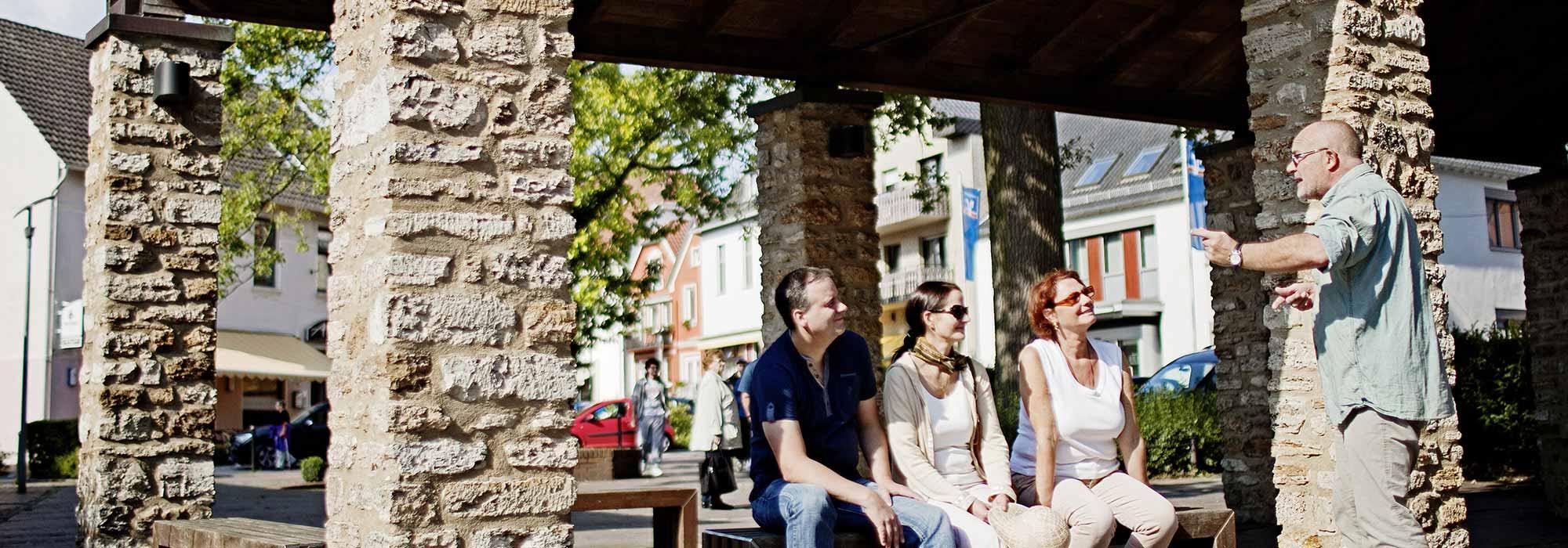 Ein Mann und zwei Frauen sitzen auf einer Bank und bekommen etwas von einem anderen Mann erklärt