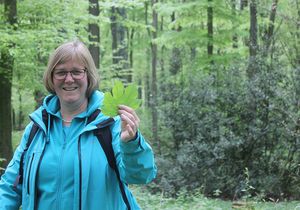 Eine Frau streckt lächelnd ein Ahornblatt im Wald in die Kamera.
